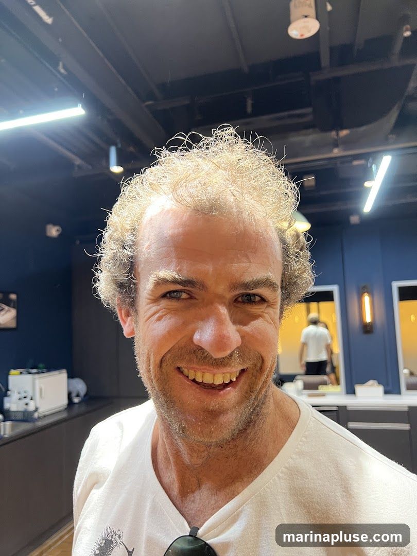A man with curly blonde hair and a beard smiles broadly at the camera. He is wearing a white t-shirt. The background appears to be a salon or barbershop, with mirrors, lighting fixtures, and a person reflected in one of the mirrors.