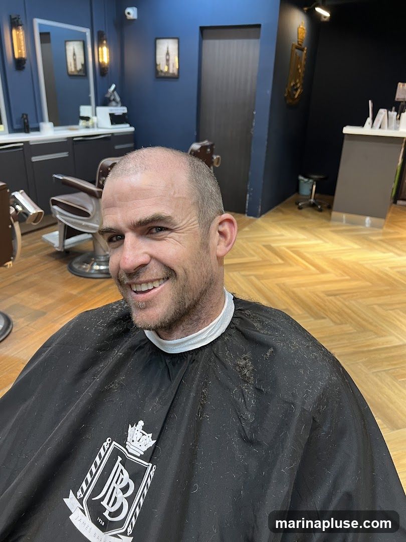 A man with a shaved head smiles at the camera, wearing a black barber cape with a logo that reads "JB 1929 JAMES BARBER". The cape is covered in hair clippings. The background shows a barber shop interior with dark blue walls, wooden herringbone flooring, vintage barber chairs, mirrors, and decorative light fixtures. A reception desk is visible in the background.