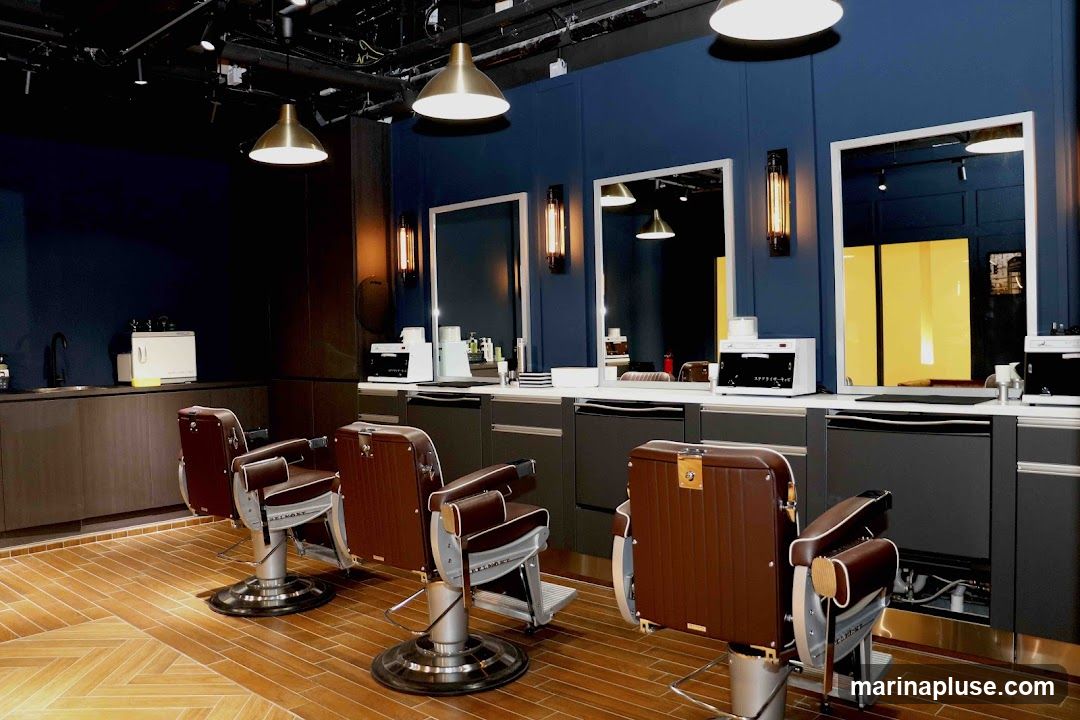A modern barbershop interior with three vintage-style barber chairs in the foreground. The chairs are upholstered in brown leather with silver accents. Behind the chairs, a dark blue wall features three large mirrors. Below the mirrors are dark gray cabinets with white countertops. Various barber equipment, including sterilizers and sinks, are visible on the countertops. The floor is made of light brown wood laid in a herringbone pattern. Industrial-style pendant lights hang from the ceiling.