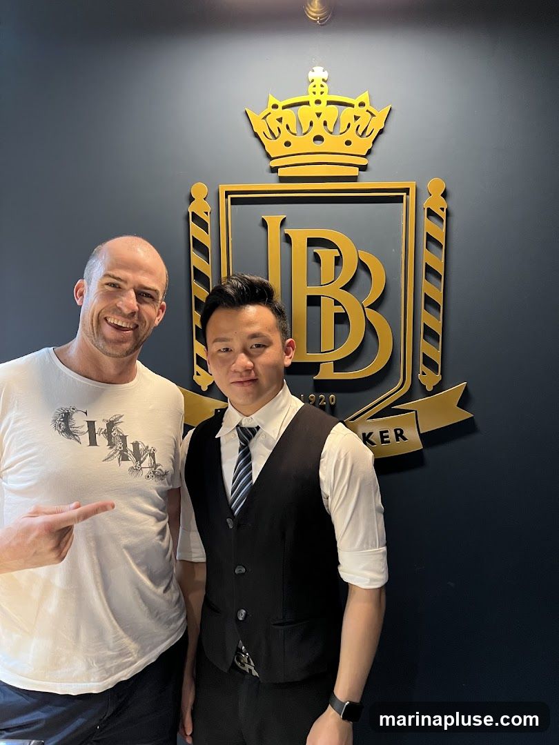 Two men are standing in front of a dark blue wall with a large, ornate gold logo. The logo features a crown at the top, a shield with the letters "JB" inside, and the word "BAKER" below it. The man on the left is wearing a white t-shirt with a graphic design and is pointing towards the logo. The man on the right is wearing a white shirt, a black vest, and a tie.