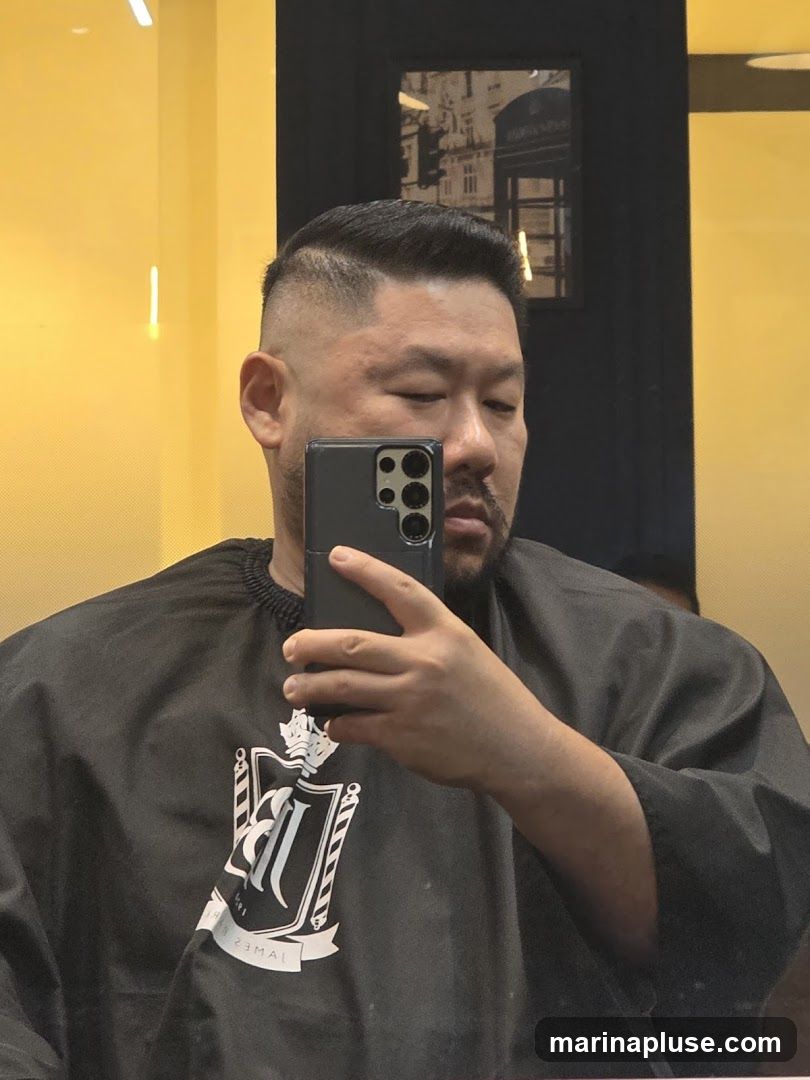A man with a fresh haircut takes a selfie in a barber shop. He is wearing a black cape with a white logo that appears to be a crest with barber poles and the name "JAMES" visible. The background is a bright yellow wall with a framed picture of a London telephone booth.