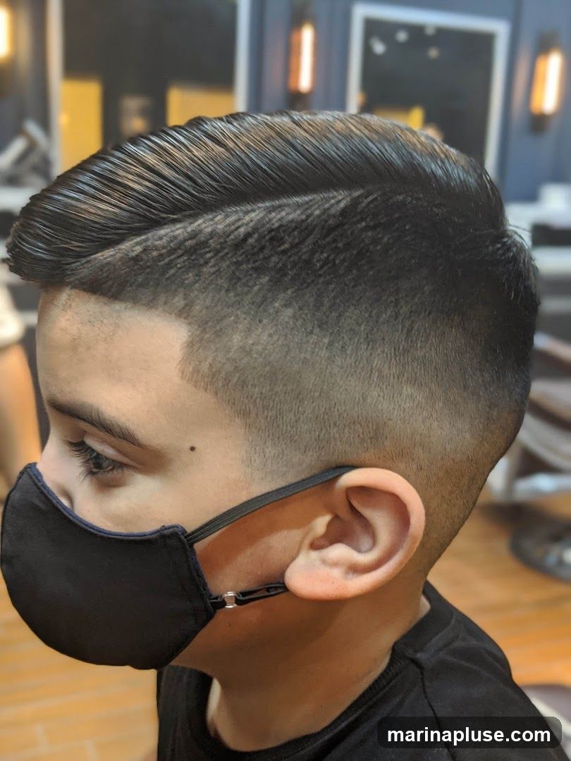 A close-up profile view of a young boy wearing a black face mask and a black t-shirt. He has a meticulously styled haircut with a sharp side part and a faded undercut. The background appears to be the interior of a barbershop, with warm lighting and dark blue walls.