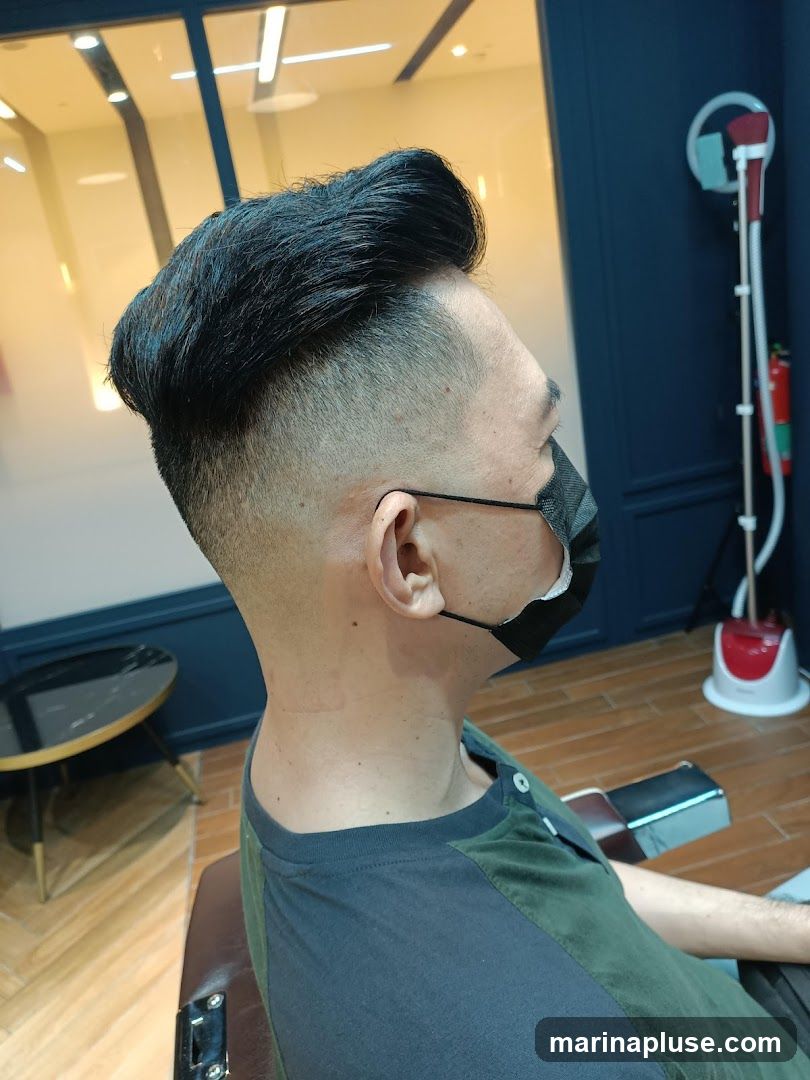 A man with a black mask on, wearing a dark green and black shirt, is sitting in a barber chair. He has a fresh haircut with a fade on the sides and styled hair on top. In the background, there is a steamer and a small round table with a marble top. The room has dark blue walls and wooden floors.