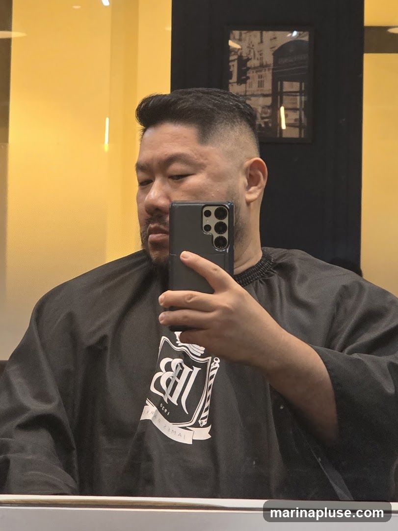 A man takes a selfie in a mirror while wearing a black barber cape with a white logo. The cape has a crest with the initials "JB" and the text "JAMES BARBER 1920". The background is a bright yellow wall and a framed picture of a London telephone booth.
