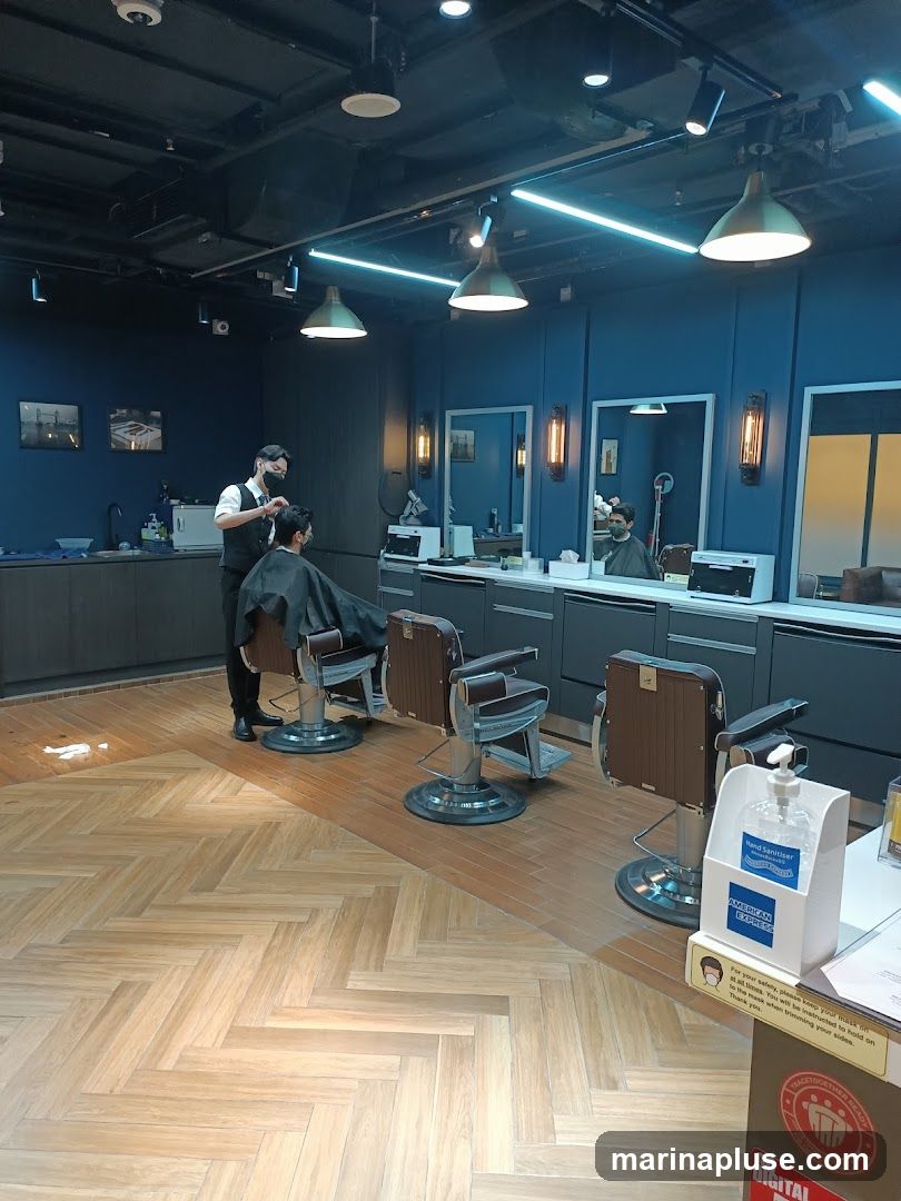 A barbershop interior with multiple barber chairs and mirrors. A barber is cutting a client's hair. The walls are dark blue, and the floor has a herringbone wood pattern. There is a hand sanitizer dispenser with an American Express logo.