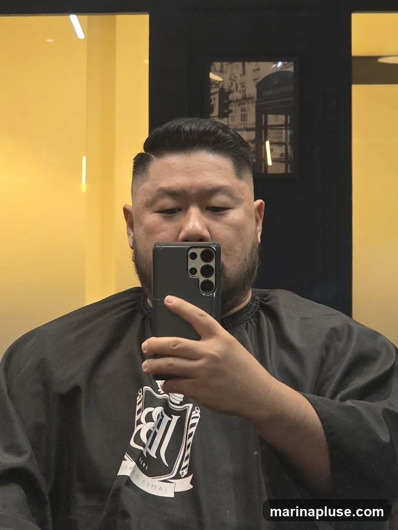 A man takes a selfie in a barber shop, wearing a black cape with a white logo that reads "JB BARBER 1930". The background is yellow and black, with a framed picture of a London telephone booth on the wall.