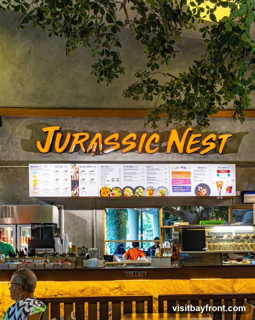 A view of the Jurassic Nest cafe counter with a large menu board above. The menu features sections for Breakfast, Local Kopi, Gourmet Coffee, Light Bites, All Day Meals, Salad, Kids Menu, Dessert, and Ice Cream. The counter area has a 'COLLECT' sign and various food items displayed.