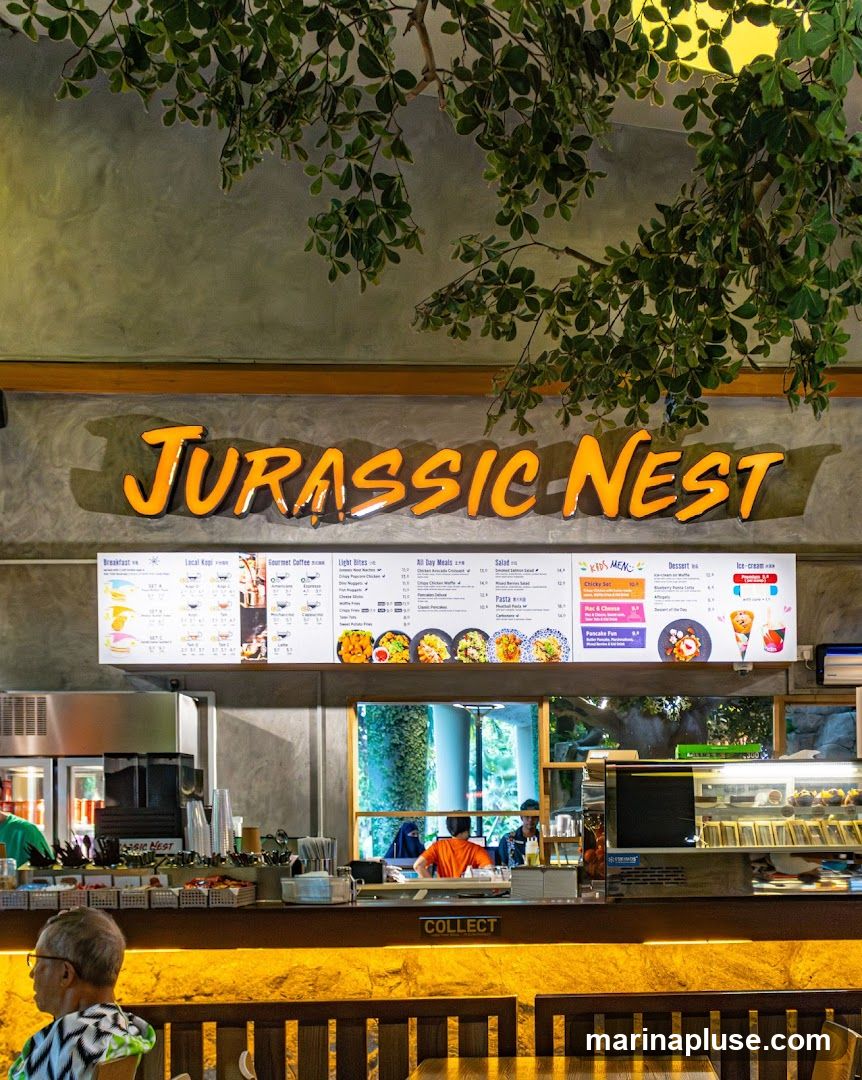 A view of the Jurassic Nest cafe counter with a large menu board above. The menu features sections for Breakfast, Local Kopi, Gourmet Coffee, Light Bites, All Day Meals, Salad, Kids Menu, Dessert, and Ice Cream. The counter area has a 'COLLECT' sign and various food items displayed.