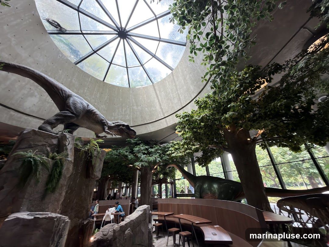 The interior of a restaurant with a jungle and dinosaur theme. A large Tyrannosaurus Rex model is perched on a rock formation on the left, while a Brachiosaurus model is visible in the background. Artificial trees and lush foliage are integrated throughout the space. Seating areas with wooden tables and chairs are arranged around the dining space. A large, circular skylight allows natural light to illuminate the area.