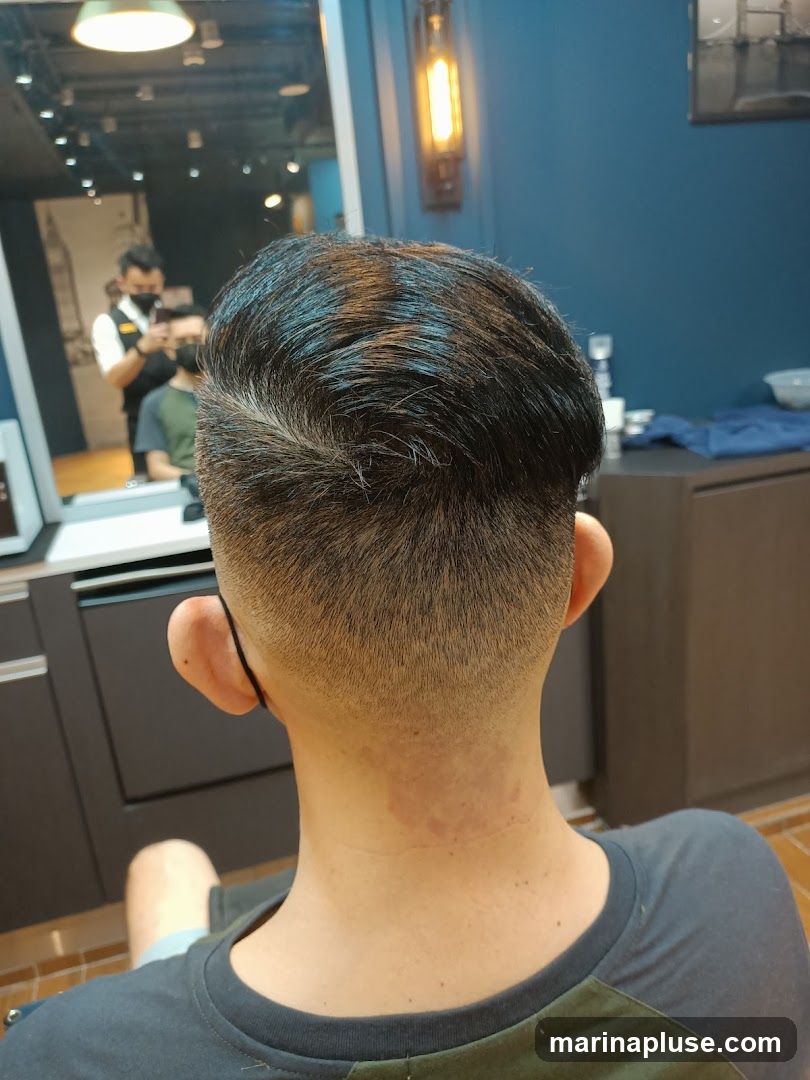 A close-up view of the back of a person's head and neck, showing a fresh haircut. The background reveals a barber shop interior with a mirror reflecting a barber wearing a mask and holding a phone, and another customer. A vintage-style light fixture hangs on a blue wall.