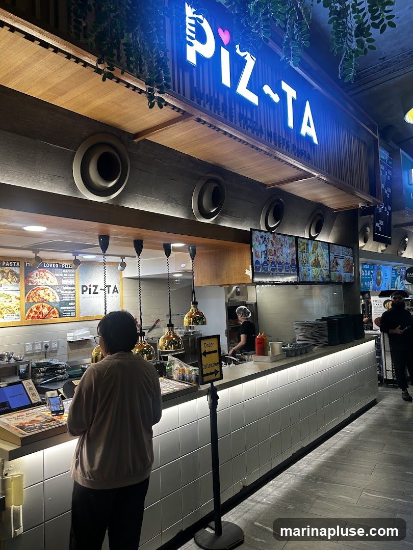A person stands at a counter in a brightly lit pizza restaurant. Above the counter, a neon sign reads "PIZ-TA". Behind the counter, staff members are preparing food. Digital screens display menu items and images of pizzas. A sign indicates "Order" and "Collection".