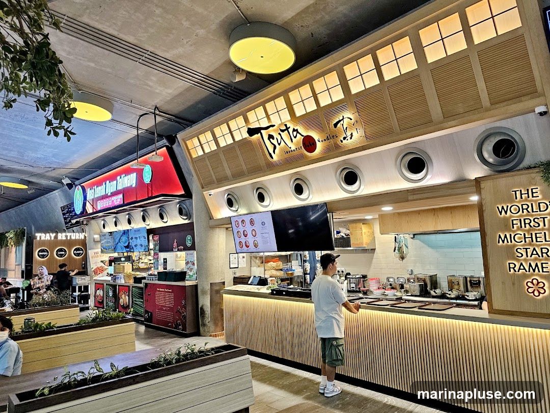 A man stands at the counter of a ramen restaurant. The counter is illuminated with warm lights and has a slatted wood facade. Above the counter, a sign reads "Tsuta Japanese Noodles." To the right, a wooden sign states "The World's First Michelin Star Ramen." In the background, a food stall with a red illuminated sign is visible, along with a "Tray Return" sign.