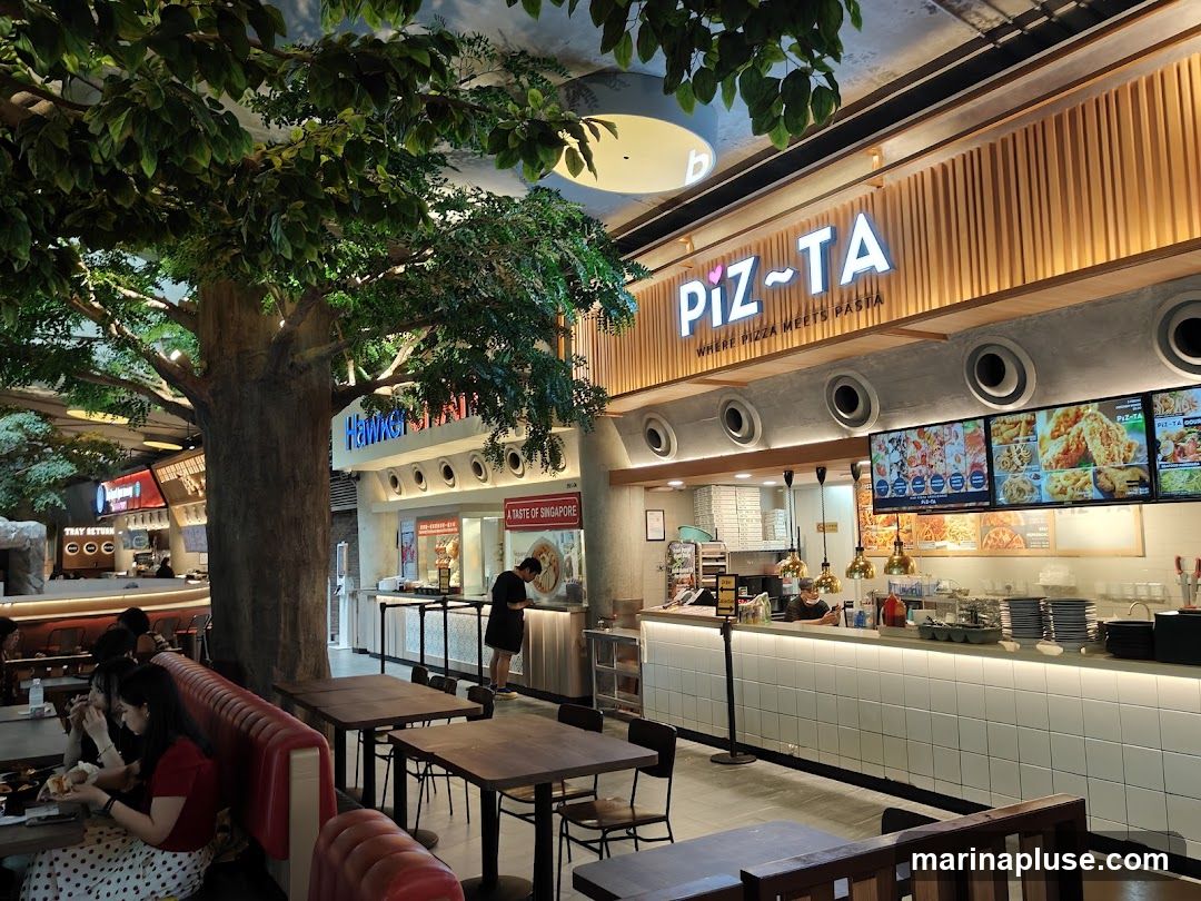 A bustling food court with various stalls, including a prominent stall named "PIZ-TA" which advertises pizza and pasta. The stall has a modern design with wooden accents and bright lighting. Digital menu boards display images of pizzas and other dishes. In the foreground, diners are seated at tables, and a large artificial tree adds a touch of greenery to the space.