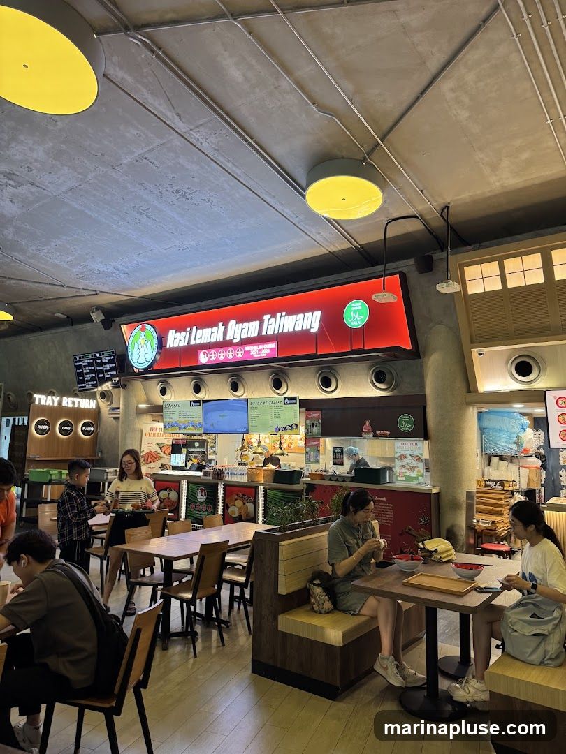A bustling food court with several stalls and dining tables. A prominent sign reads "Nasi Lemak Ayam Taliwang" with a Halal certification. People are seen eating at tables and ordering food. There are also signs for "Tray Return" and various menu boards.