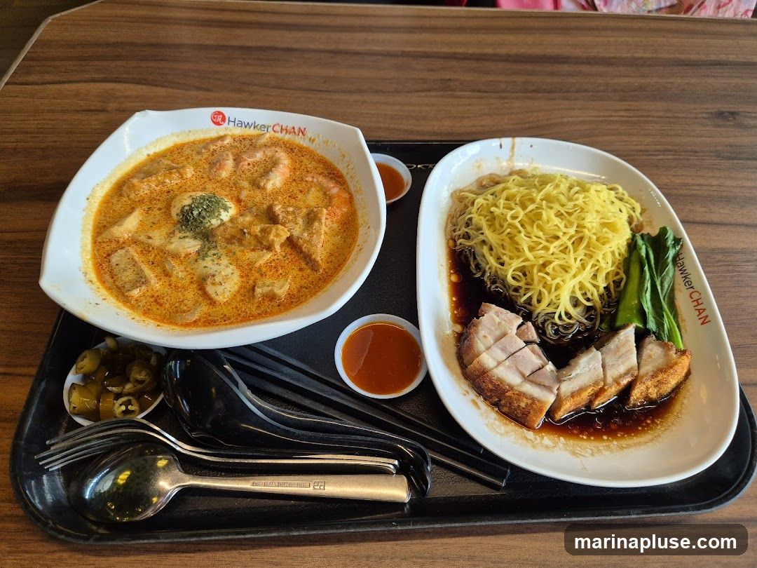 A tray with two bowls of food and cutlery. One bowl contains laksa with shrimp and tofu. The other bowl contains noodles with roasted pork and greens. There are also small dishes of chili sauce and pickled chilies. The bowls have the Hawker Chan logo.