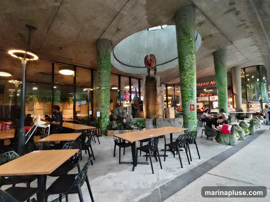 An indoor cafe with a dinosaur theme. Tables and chairs are set up for dining. A large dinosaur statue is visible in the background, along with a sign that reads "JURASSIC NE... JN Cafe". The pillars are decorated to look like they are covered in moss.