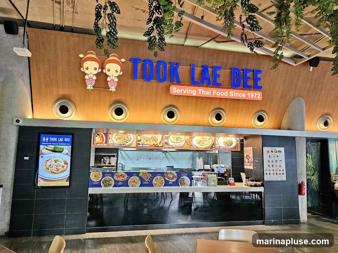 A food stall with the name "TOOK LAE DEE" prominently displayed in blue letters, along with the tagline "Serving Thai Food Since 1972". The stall features a display of various Thai dishes with their names and images, including Tom Yum Soup, Spicy Venicelli Salad, and Prawn Tom Yum. There is also a digital menu board showing a dish of Som Tum Thai Green Papaya Salad. The stall is decorated with cartoon characters and hanging plants.