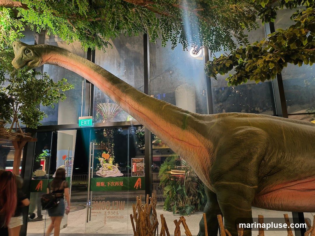 A large dinosaur model is prominently displayed in front of a glass-enclosed entrance. An "EXIT" sign is visible above the doorway. Inside, there are posters and decorations, including what appears to be a display with a dinosaur figurine. Several people are seen walking near the entrance. The scene is enhanced with artificial trees and foliage, creating a prehistoric ambiance.