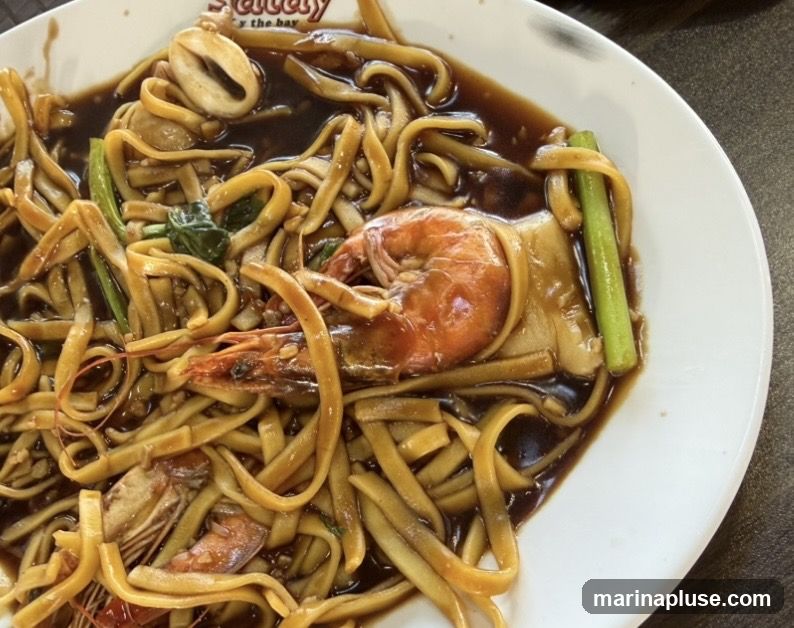 A close-up shot of a plate of noodles with seafood. The noodles are thick and flat, coated in a dark, savory sauce. Several large shrimp and pieces of squid are visible, along with some green vegetables. The dish is served on a white plate with a partial view of a logo that reads "Seafood the bay" at the top.