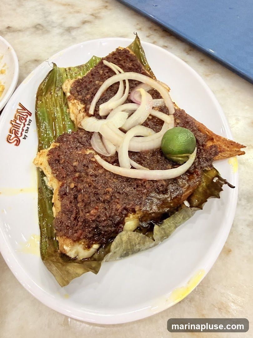 A white plate holds a fish dish served on a banana leaf. The fish is topped with a dark, textured sauce and garnished with sliced white onions and a lime wedge. The plate has a logo that reads "Satay by the bay".