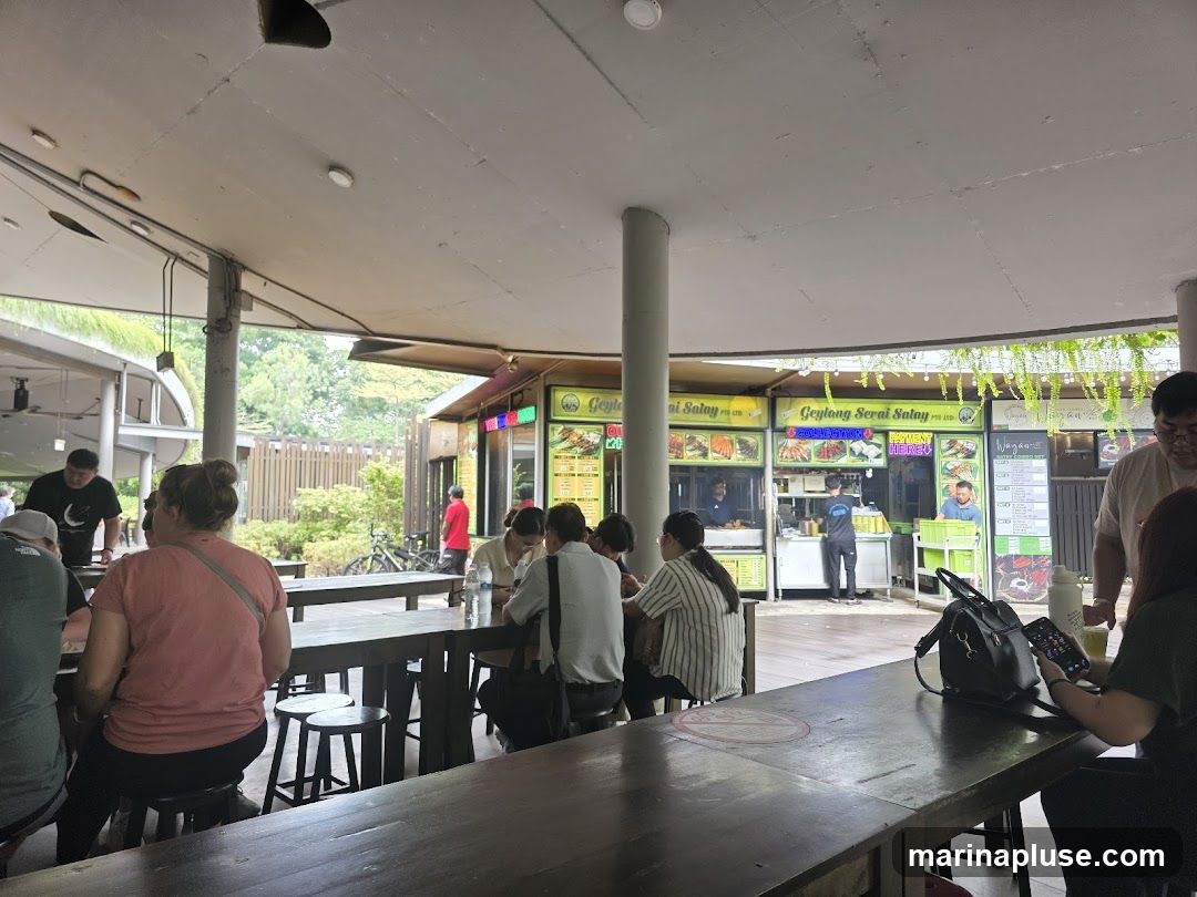 People are seated at long wooden tables under a covered outdoor area. In the background, several food stalls are visible, with signage indicating "Geylang Serai Satay" and "Vegan Briyani". Staff members are seen working at the stalls, and customers are interacting with them. The area appears to be a casual dining spot with a relaxed atmosphere.