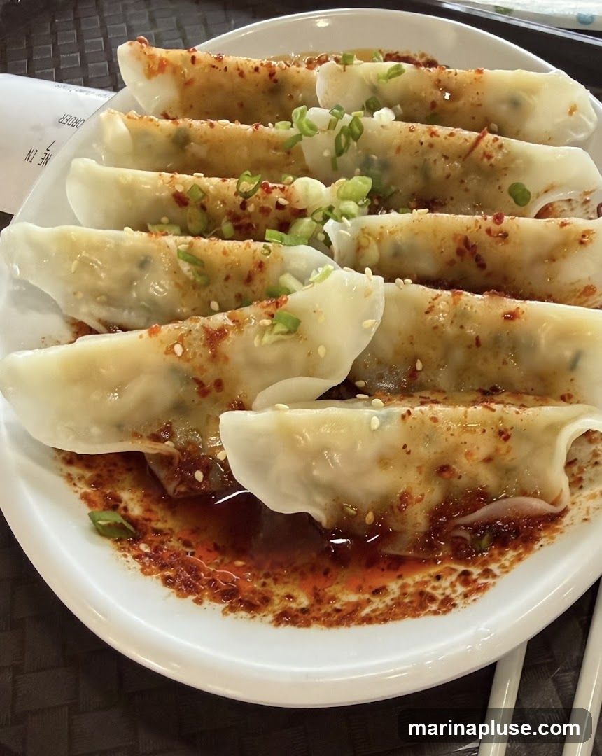 A plate of dumplings, likely pan-fried or boiled, is served with a savory sauce and garnished with chopped green onions and sesame seeds. The dumplings are arranged in rows on a white plate, with a dark, textured placemat visible beneath. A receipt or order slip is partially visible in the upper left corner.