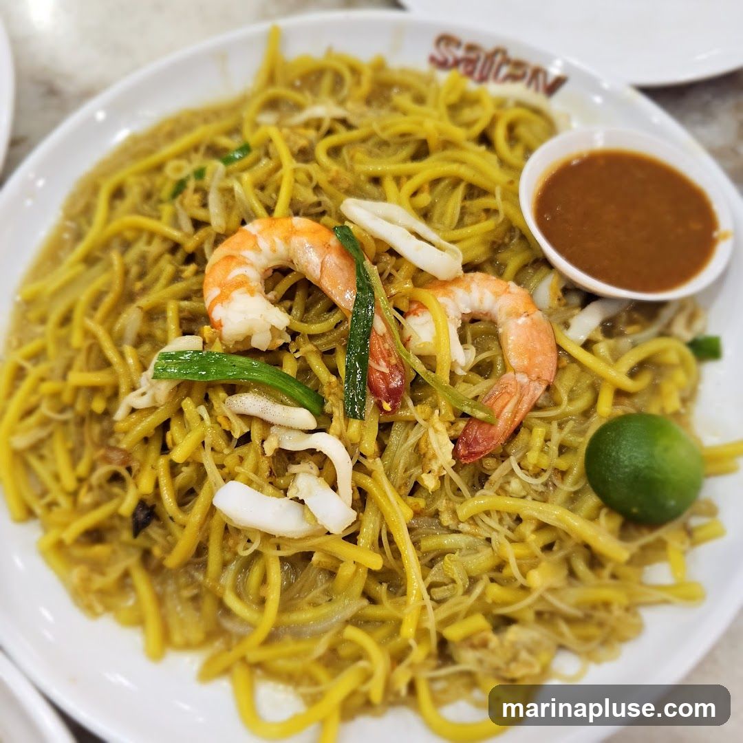A plate of yellow noodles with shrimp, squid, and green onions, served with a side of dipping sauce and a lime wedge. The word "Satey" is visible on the rim of the plate.