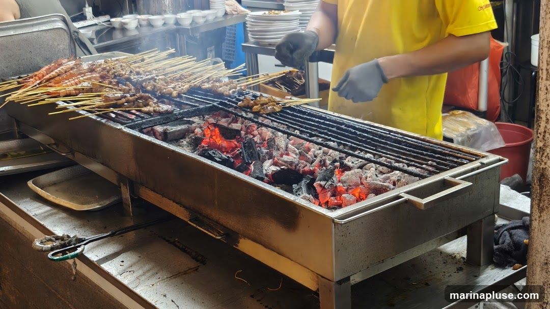 A person in a yellow shirt and grey gloves is grilling skewers of meat and shrimp over hot coals. The skewers are arranged on a metal grill. In the background, there are stacks of plates and bowls, and other food preparation items.