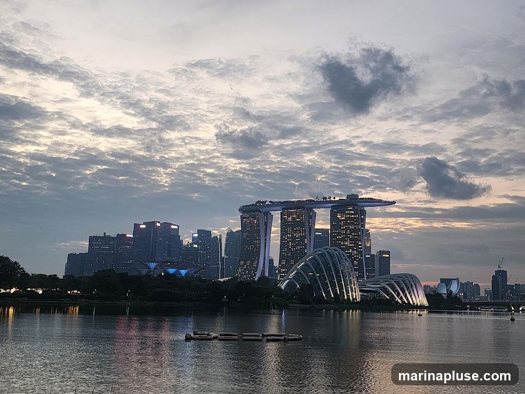 A panoramic view of the Singapore skyline at dusk, featuring the iconic Marina Bay Sands hotel, Gardens by the Bay, and other skyscrapers. The sky is filled with dramatic clouds, and the city lights are beginning to illuminate the buildings. The calm water in the foreground reflects the city lights and the sky.