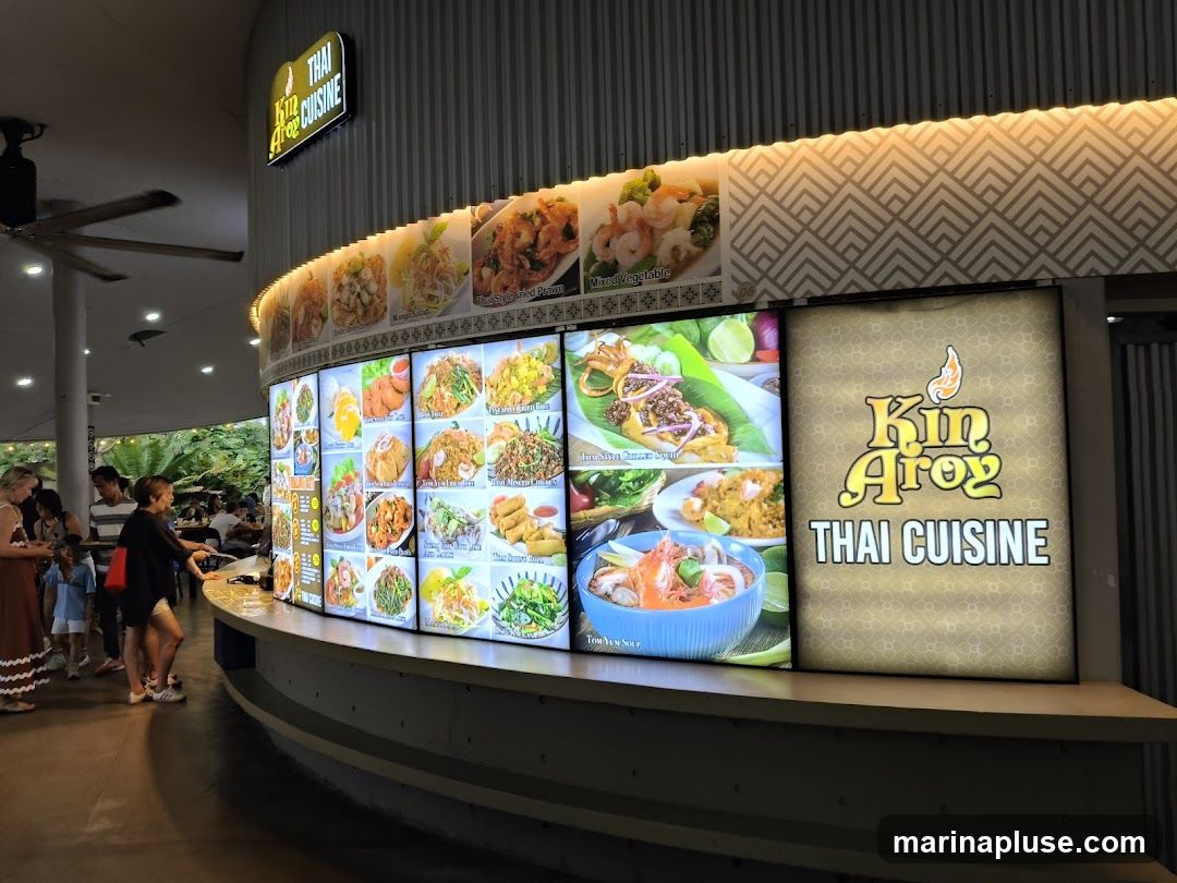 A brightly lit display of various Thai dishes is shown, with a prominent sign for "Kin Aroy Thai Cuisine" on the right. People are seen ordering food at the counter. The display features images of dishes like Pad Thai, Tom Yum Soup, and various stir-fries.