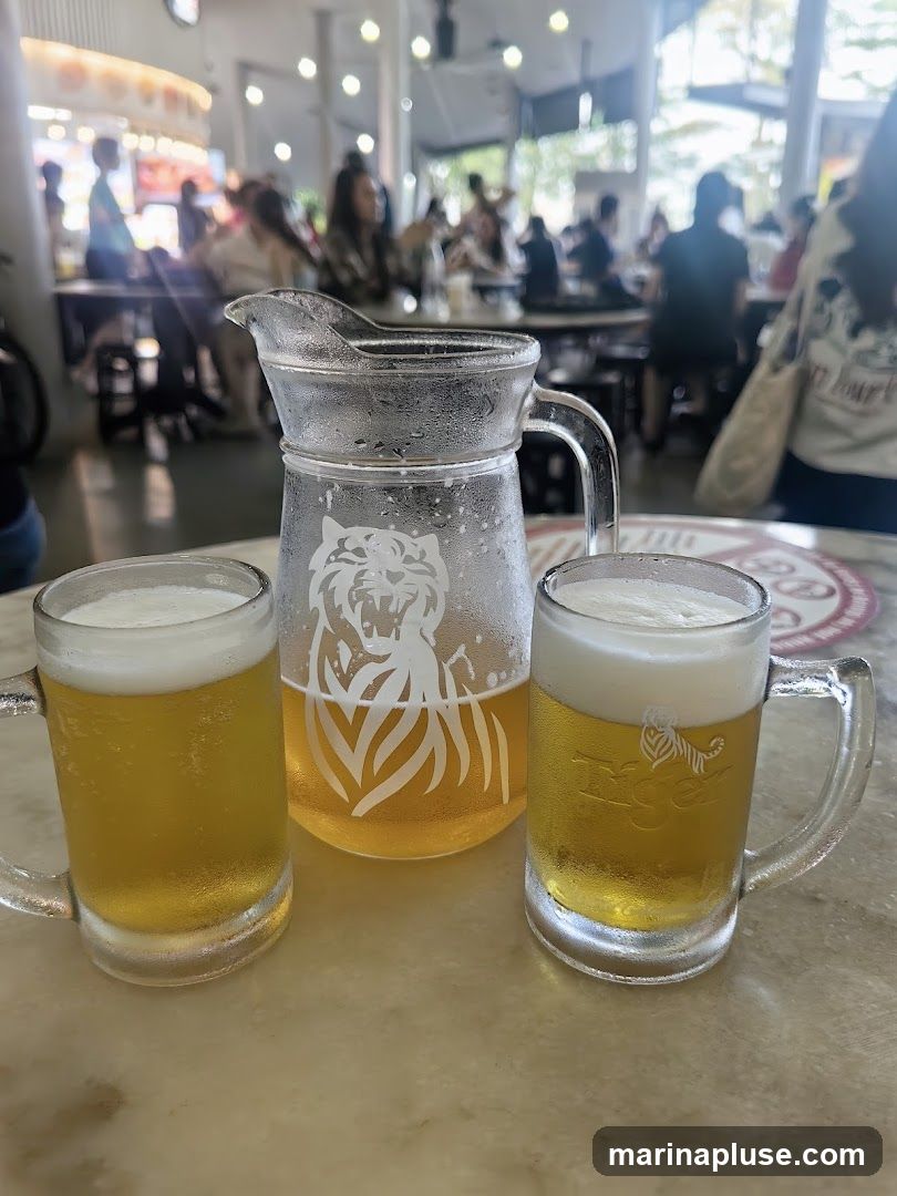 Two mugs of beer and a pitcher of beer are on a marble table. The pitcher has a tiger logo etched into it. In the background, people are seated at tables in a brightly lit indoor/outdoor space.