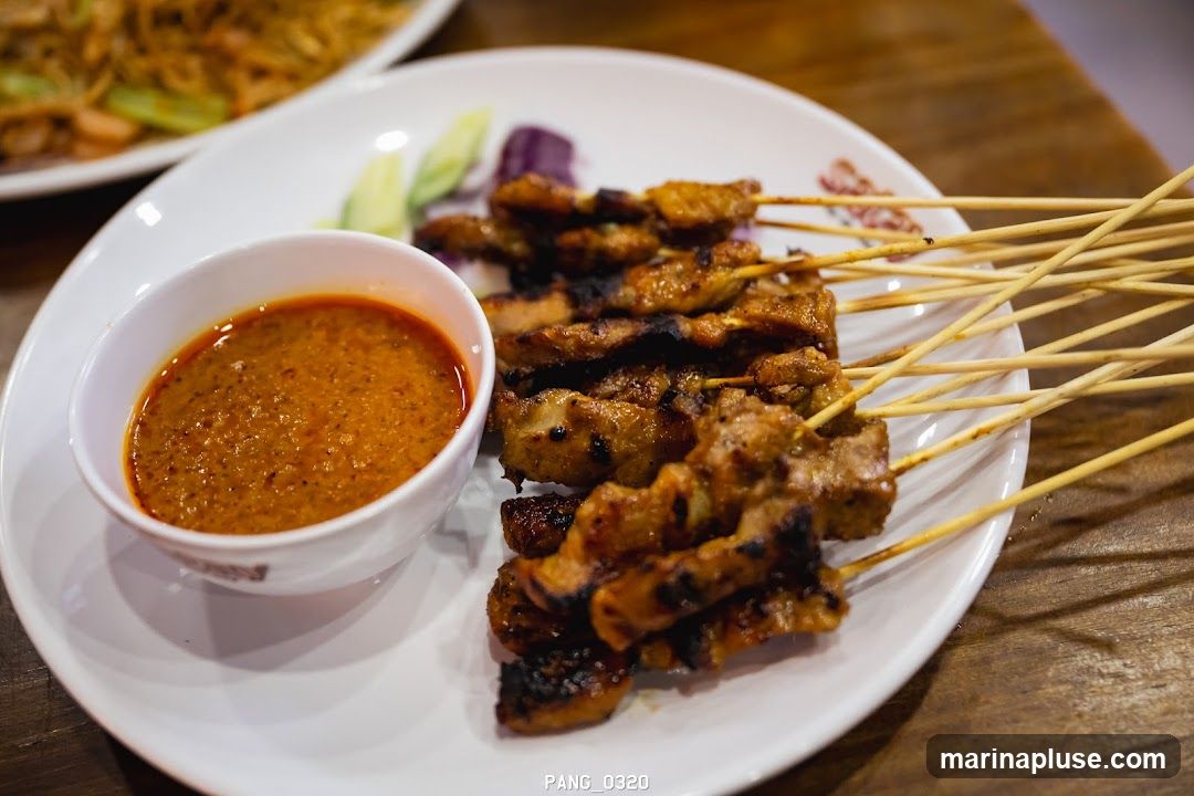 A close-up shot of a plate of chicken satay skewers served with a side of peanut sauce. The satay appears grilled and slightly charred, with a rich, textured peanut sauce in a white bowl. Cucumber slices and red onion are visible as garnishes. In the background, another plate of food, possibly noodles, is out of focus. The image is taken from a slightly elevated angle, focusing on the food.