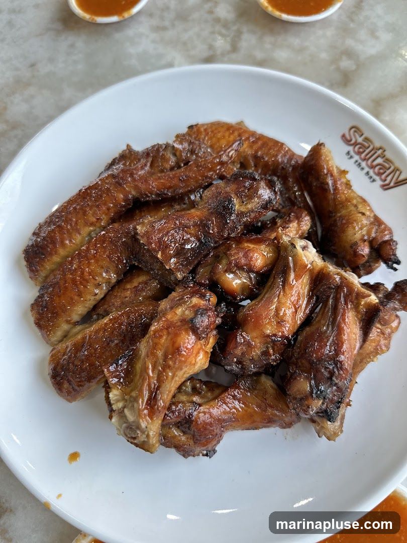 A plate of glistening, golden-brown chicken wings is presented on a white plate. The wings appear to be roasted or grilled, with some char marks visible, suggesting a savory and slightly smoky flavor. The plate has a logo that reads "Satay by the bay". Two small dipping bowls with orange sauce are visible in the background.