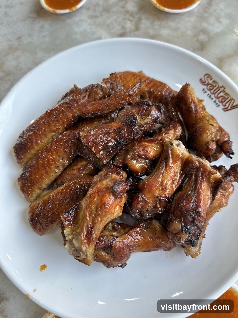 A plate of glistening, golden-brown chicken wings is presented on a white plate. The wings appear to be roasted or grilled, with some char marks visible, suggesting a savory and slightly smoky flavor. The plate has a logo that reads "Satay by the bay". Two small dipping bowls with orange sauce are visible in the background.