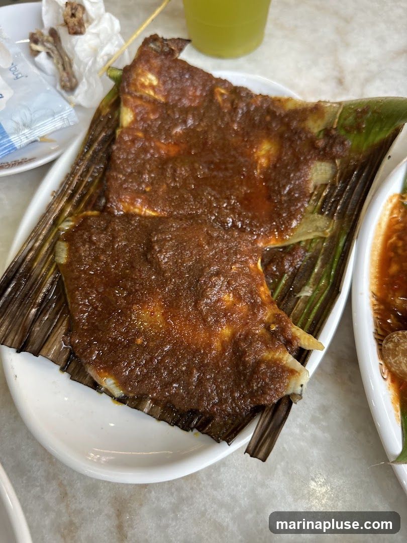 Two pieces of grilled fish, covered in a thick, reddish-brown spicy sauce, are served on a bed of grilled banana leaves. The fish appears flaky and moist. A side dish with a similar sauce and a green beverage are also visible. A small plate with remnants of food and a wet wipe is in the background.