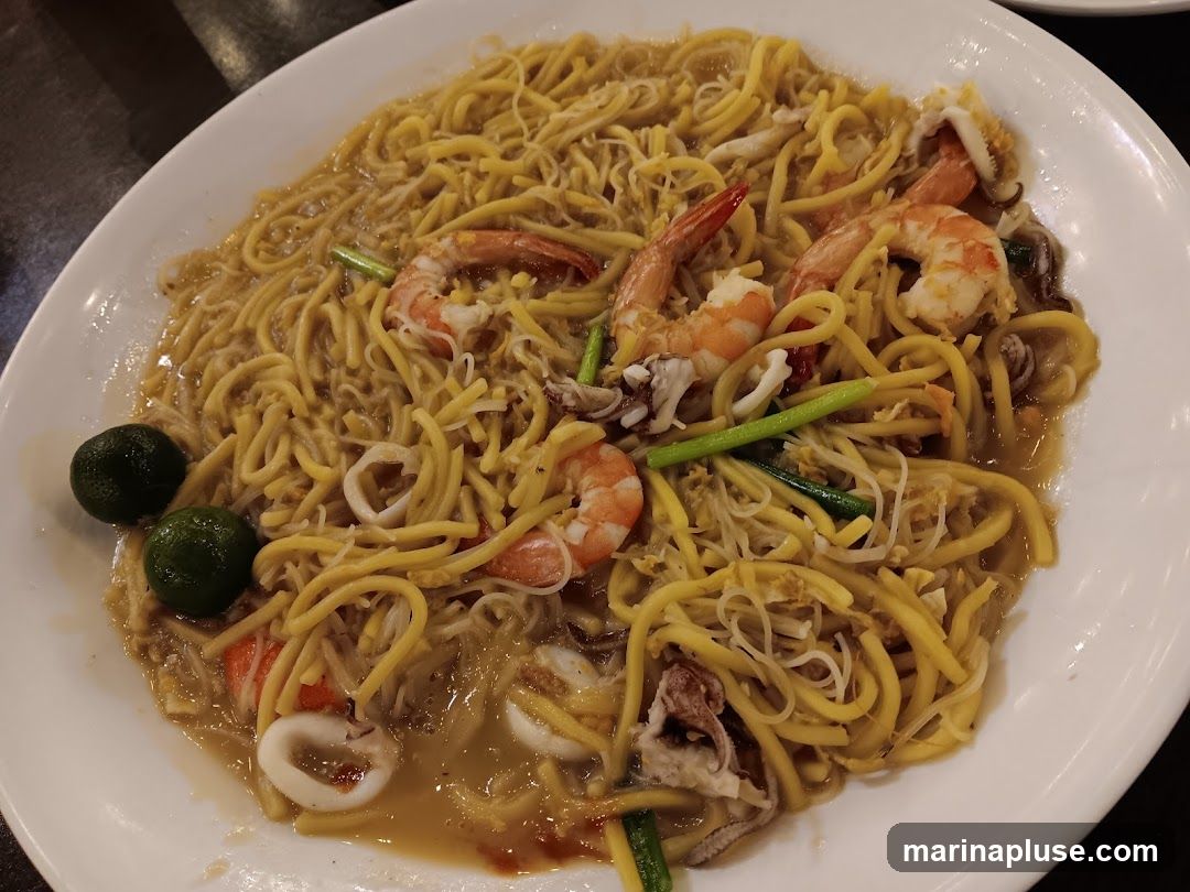 A close-up shot of a plate of seafood noodles, featuring yellow noodles mixed with shrimp, squid rings, and bean sprouts, garnished with green onions and lime wedges. The dish is served on a white oval plate.