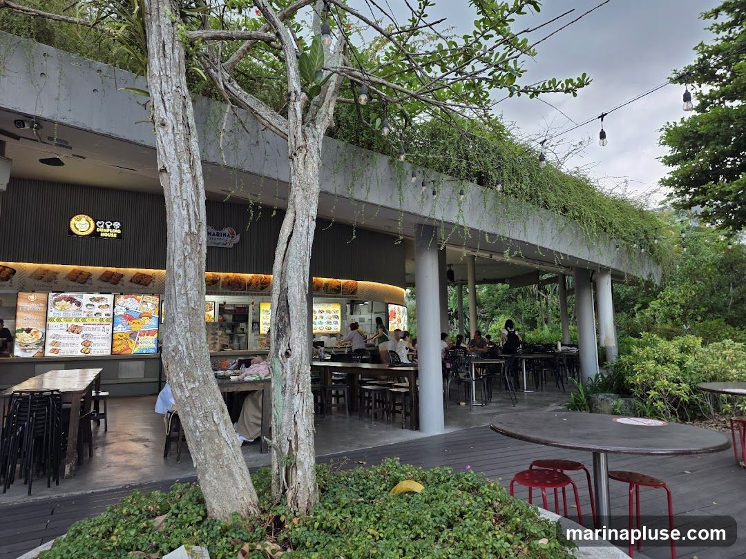 An outdoor food court with several stalls and seating areas. Lush greenery covers the upper level of the building, and trees are in the foreground. String lights are visible overhead. People are seated at tables, and food is displayed at the stalls.