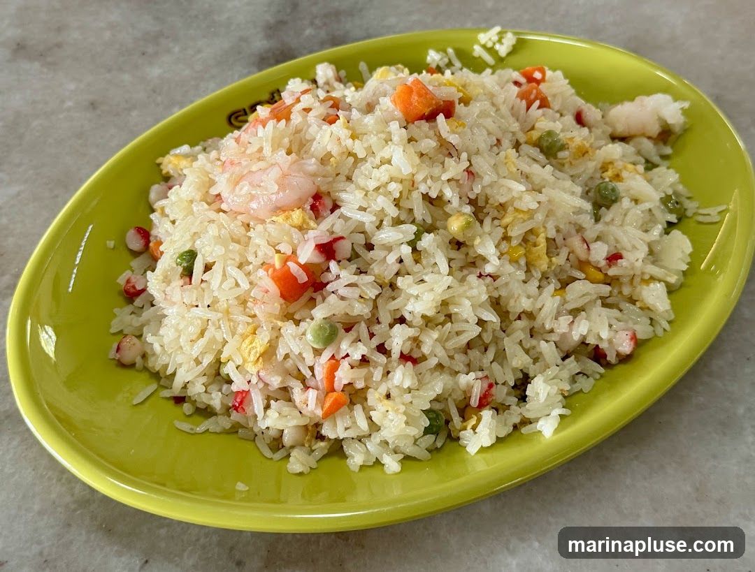 A close-up shot of a plate of shrimp fried rice. The rice is mixed with peas, carrots, scrambled egg, and pieces of shrimp and imitation crab. The dish is served on a bright green oval plate.