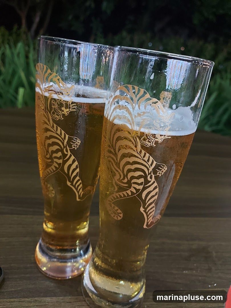 Two tall glasses filled with golden beer and a frothy head. The glasses are decorated with a stylized golden tiger design. They are placed on a dark wooden surface, with a blurred background of greenery and lights.