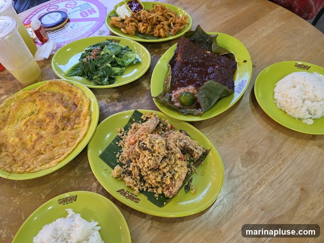 A variety of dishes are spread across a wooden table. In the foreground, there are two plates of white rice, one partially visible. To the left, a large omelet is served on a bright green plate. In the center, a dish of fried food with a crumbly coating is presented on a banana leaf. Behind this, a piece of fish in a dark sauce, wrapped in leaves, sits on a green plate, with a lime wedge on top. To the right, a plate of white rice is visible. Further back, a plate of fried calamari with dipping sauces is present. A plate of green vegetables is also on the table. Two plastic cups with a light-colored beverage are on the far left. The plates and cups have a logo that appears to say "Satay by the Bay".