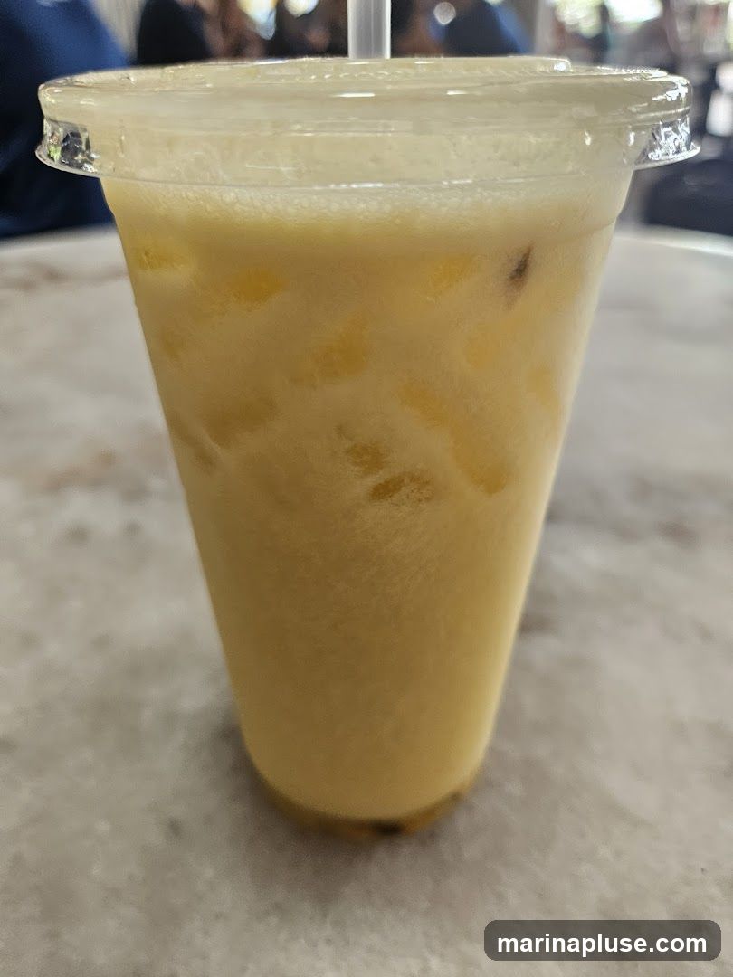 A close-up shot of a yellow, frothy iced beverage in a clear plastic cup with a straw. The drink appears to have pulp or small pieces of fruit mixed in. The cup is on a speckled, light-colored surface, possibly a table. In the blurred background, people and an outdoor setting are faintly visible.