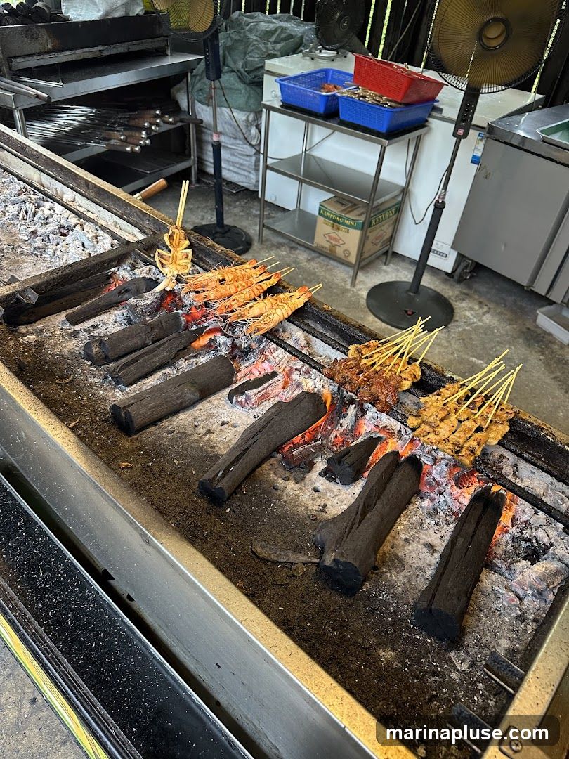 A close-up view of a charcoal grill with various skewers of food being cooked. In the foreground, several large prawns are grilling, and a whole squid is skewered and positioned over the coals. To the right, rows of satay skewers, appearing to be chicken or beef, are also being grilled. The grill is filled with glowing charcoal and ash. In the background, stainless steel shelves hold blue and red plastic containers, and industrial fans are visible.