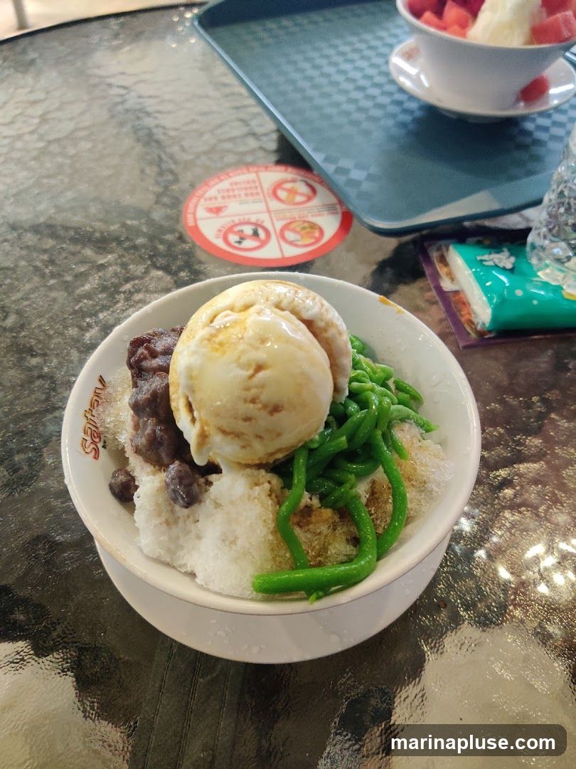 A bowl of shaved ice dessert topped with a scoop of ice cream, red beans, and green jelly noodles. The bowl has a logo that reads "Safa" on its rim. A tray with a bowl of fruit and ice is visible in the background, along with a packet of tissues. A circular red and white sign with pictograms is on the table.