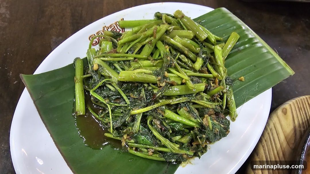 A close-up shot of stir-fried water spinach, also known as kangkong, served on a white plate lined with a banana leaf. The dish appears to be seasoned with garlic and chili, and is glistening with sauce. The plate has a subtle embossed pattern around the rim, with the word "SATAY" visible in stylized lettering.
