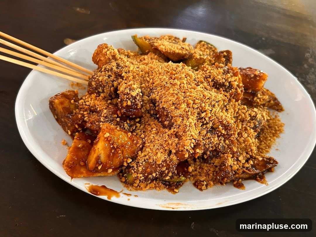 A white plate filled with Rojak, a Malaysian fruit and vegetable salad, topped with crushed peanuts. The Rojak appears to be a mix of fried dough, fruits, and vegetables coated in a dark, savory sauce. Wooden skewers are visible on the left side of the plate. The dish is presented on a dark, textured surface.