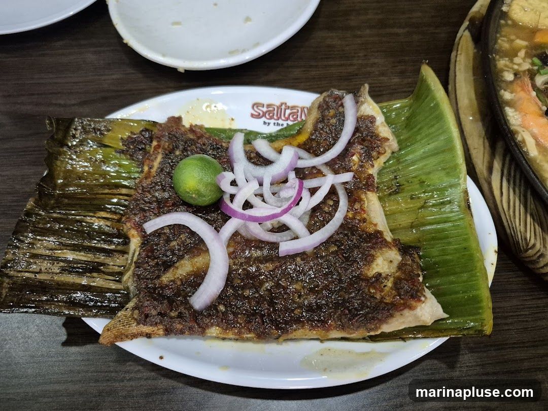 A close-up shot of a piece of grilled fish served on a banana leaf and a white plate. The fish is covered in a dark, spiced marinade and topped with sliced red onions and a lime wedge. In the background, another dish with shrimp and vegetables in a broth is visible. The plate has a logo that reads "Saitan by the Sea".