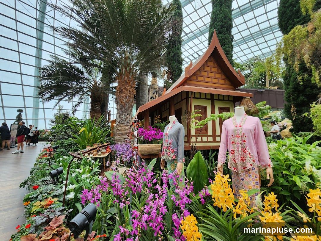 A vibrant display of orchids and other tropical plants surrounds a miniature traditional wooden house. Two mannequins are dressed in traditional attire, one in pink and the other in grey, both adorned with intricate embroidery. Palm trees and other lush greenery fill the background, all enclosed within a glass-domed structure.