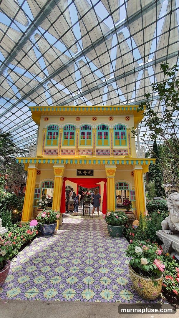 A vibrant yellow building facade with colorful window frames and decorative elements is set against a backdrop of a glass-domed structure. Red drapes hang at the entrance, and people are seen entering. The ground is tiled with a patterned design, and potted flowers and greenery surround the structure, including a stone lion statue on the right.