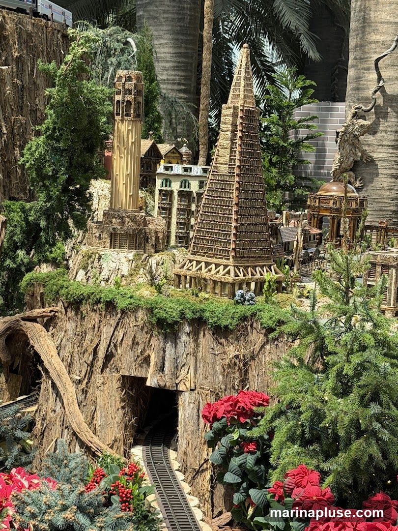 A detailed miniature village scene featuring various architectural styles, including a tall, slender tower and a pyramid-shaped building. Train tracks run through a tunnel carved into a rocky, tree-covered landscape. Red poinsettias and evergreen trees add a festive touch to the diorama. A train is visible on the tracks in the upper left corner.