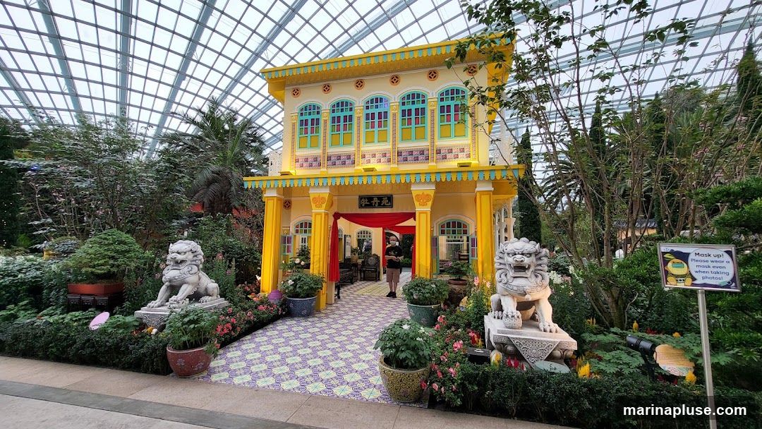 A decorative yellow building facade, resembling traditional architecture, is set within a lush greenhouse environment. The facade features colorful windows and intricate detailing. Flanking the entrance are two stone lion statues. Red drapes hang at the entrance, and a person wearing a mask is visible inside. The ground is tiled with a patterned design. A sign reminding visitors to wear masks is also present.